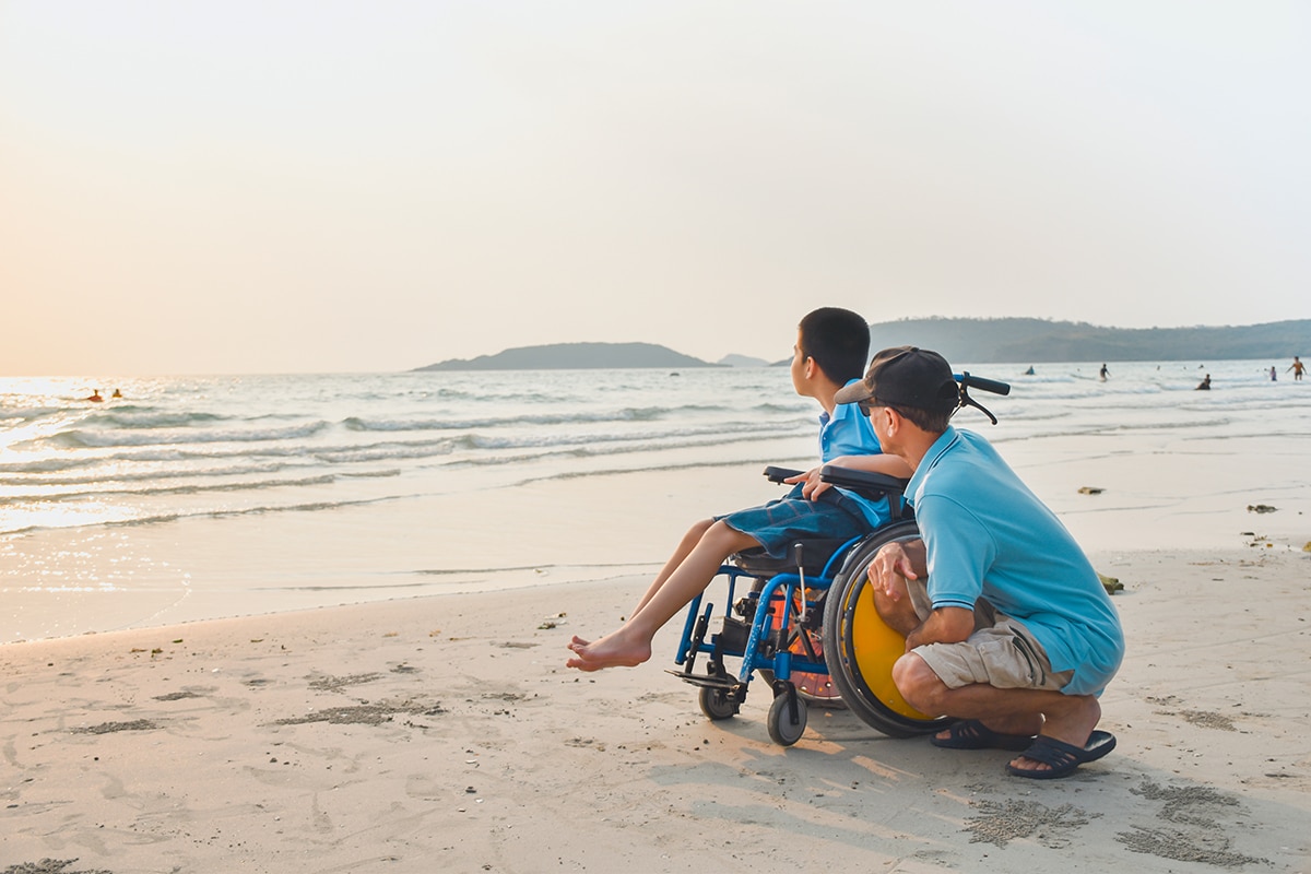 Child on wheelchair and his dad on the beach at sunset.
